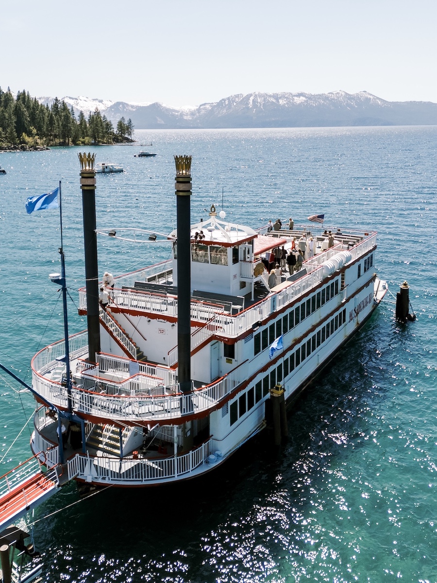 A large paddlewheel steamboat docked at a pier on a lake, with people on the upper deck and mountains in the background.