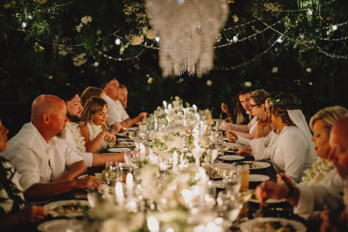 A group of people dressed in white sit around a long, elegantly decorated dinner table with candles and flowers, dining under string lights.