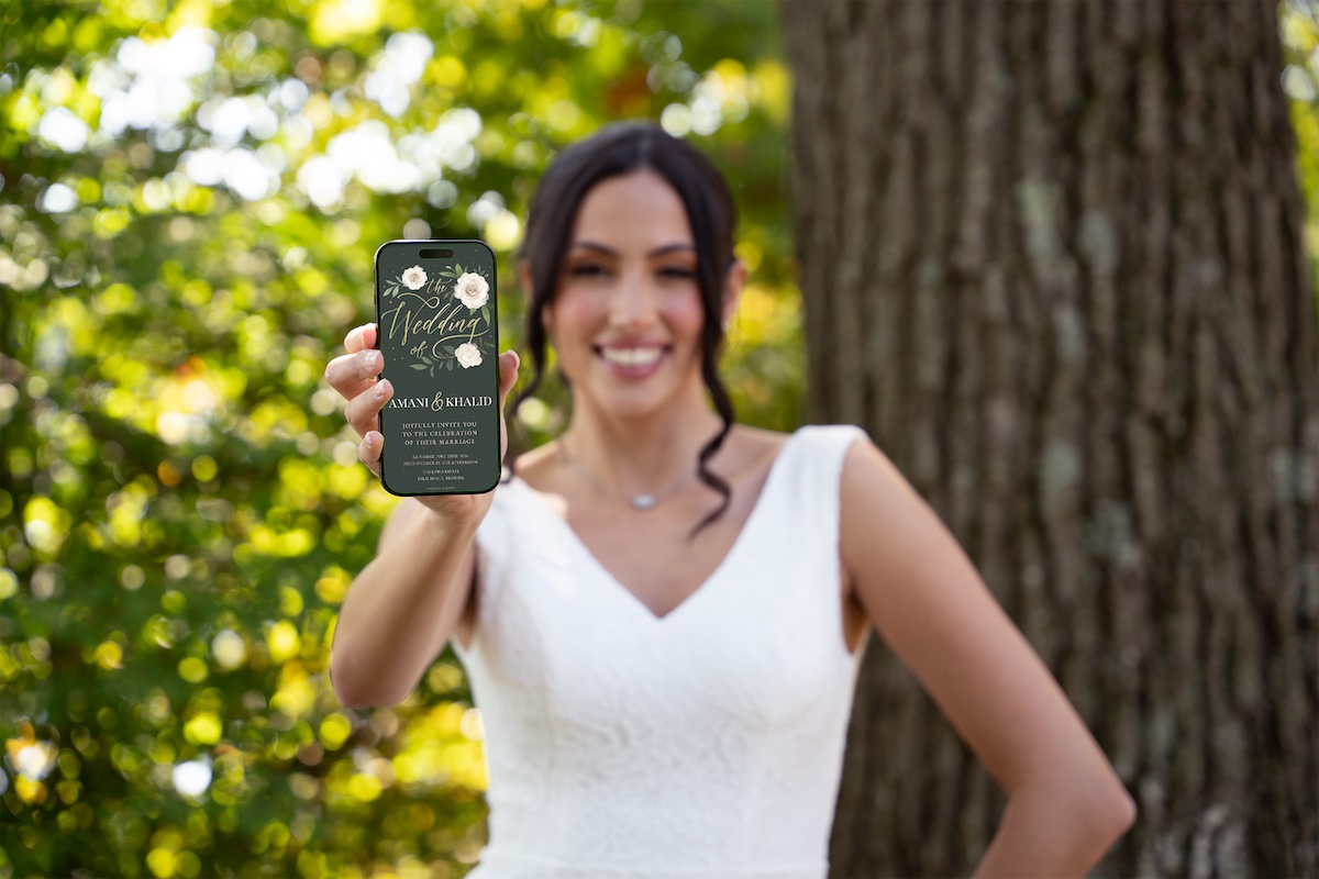 A woman in a white dress stands outdoors, smiling and holding a bottle of beverage toward the camera with a tree and greenery in the background.