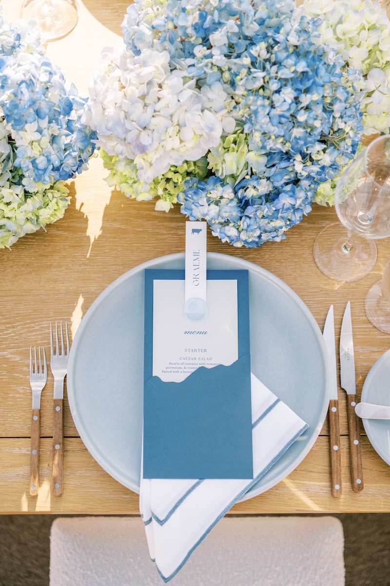 A table setting with a blue plate, white napkin, blue menu card, wooden cutlery, wine glasses, and blue hydrangea floral arrangements as the centerpiece.