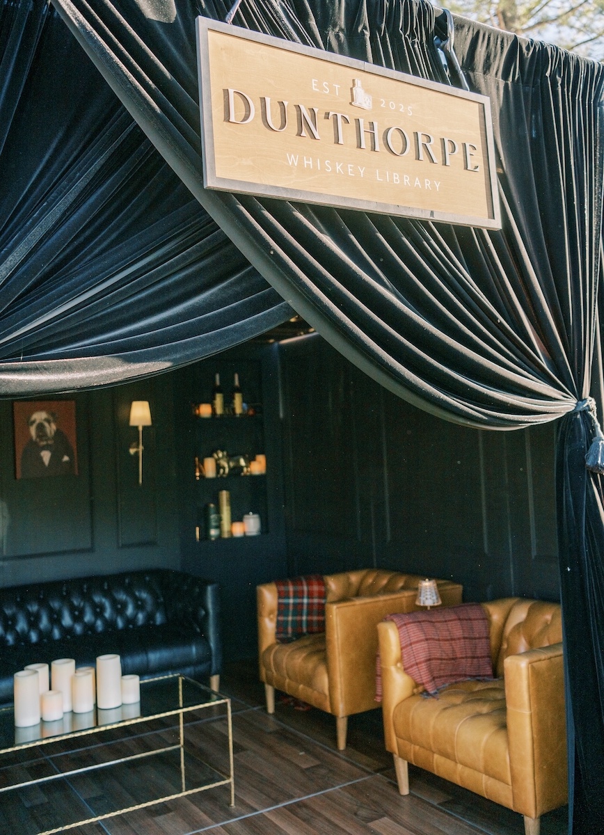 A lounge area with a black sofa, two tan armchairs, a glass coffee table with candles, shelves with bottles, and a Dunthorpe Whiskey Library sign overhead.