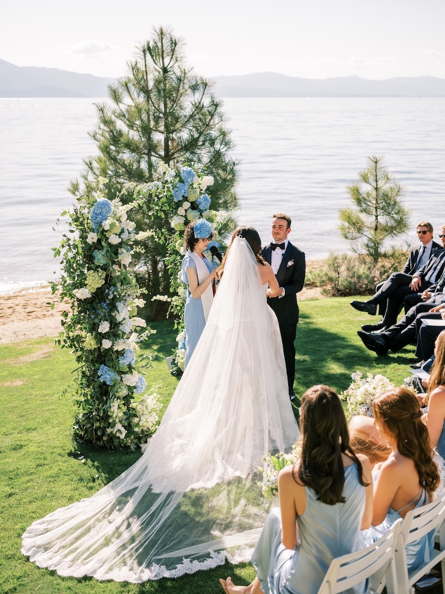 A bride and groom stand by a floral arch at an outdoor lakeside wedding ceremony, surrounded by guests seated on white chairs.