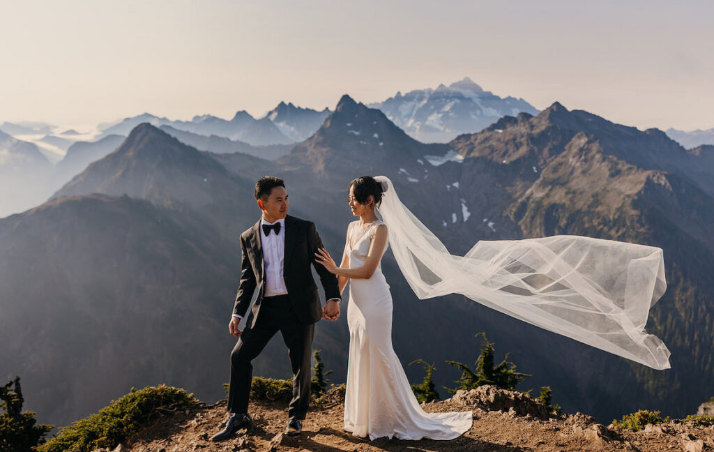 A bride and groom stand on a mountain ledge, holding hands, with mountains in the background and the bride's veil flowing in the wind.