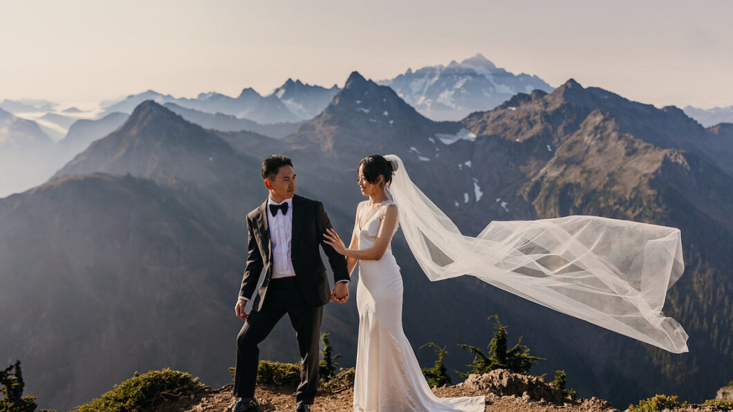 A bride and groom stand on a mountain ledge, holding hands, with mountains in the background and the bride's veil flowing in the wind.