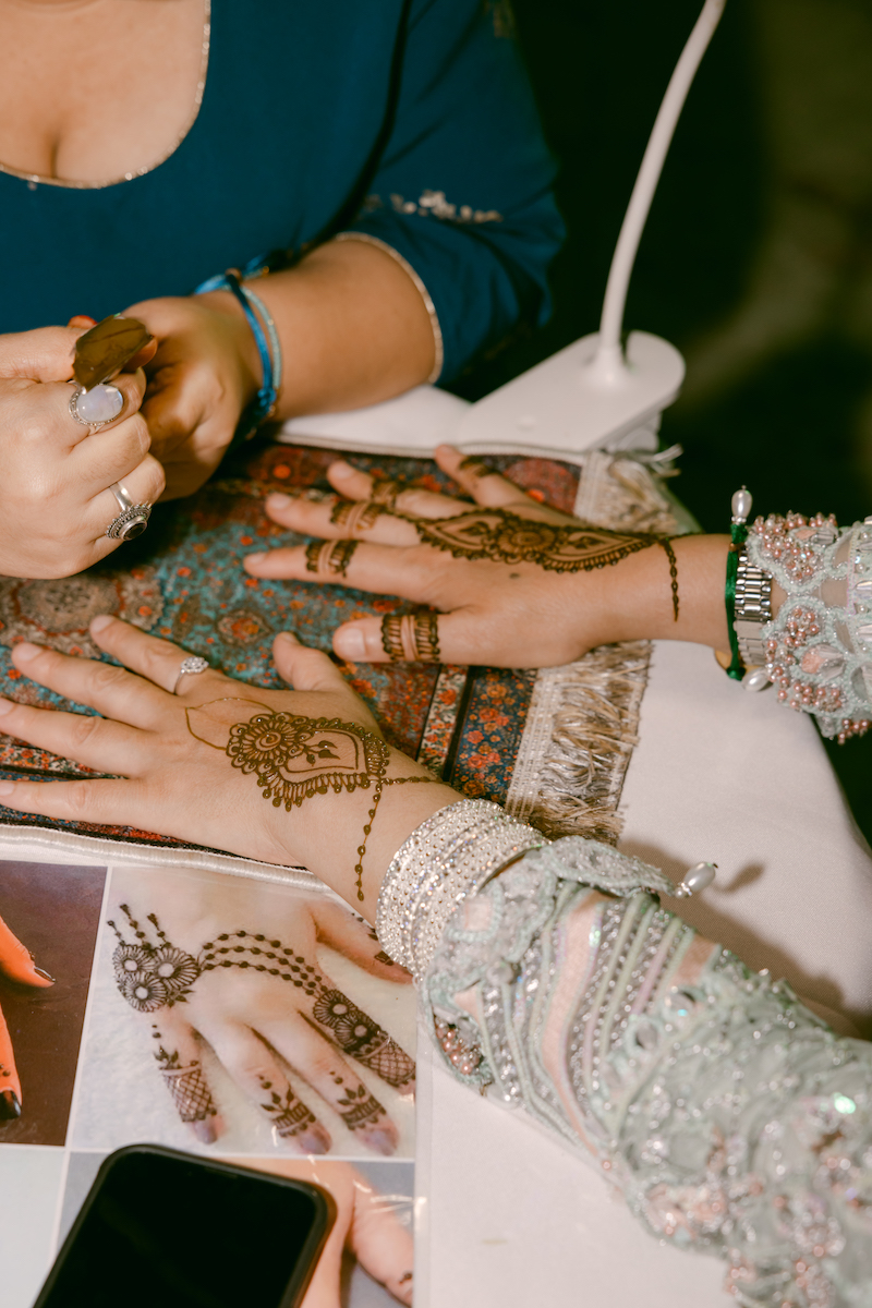 Person applying intricate henna designs to another person's hands at a table, with reference photos and a phone nearby.