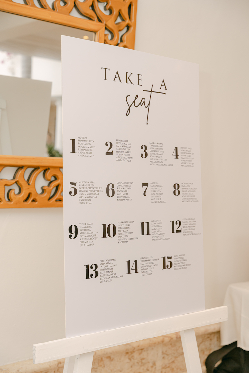 A large white seating chart with the heading "Take a seat" lists tables numbered 1 to 14, each with names underneath, displayed on an easel near a decorative mirror.