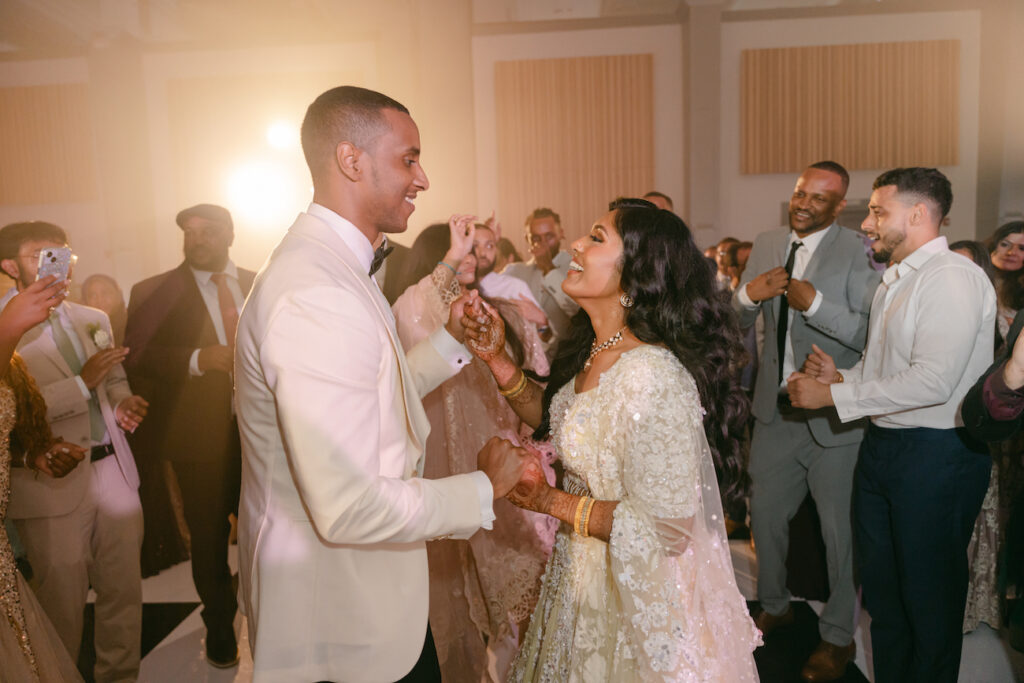 A couple in formal attire dances joyfully at a wedding reception, surrounded by smiling guests on the dance floor.