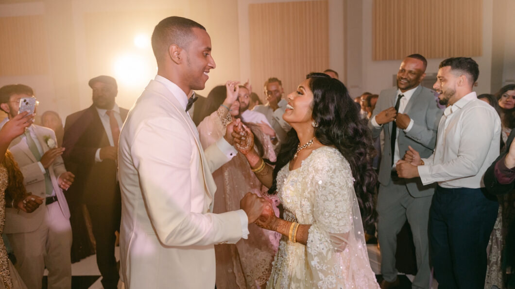 A couple in formal attire dances joyfully at a wedding reception, surrounded by smiling guests on the dance floor.