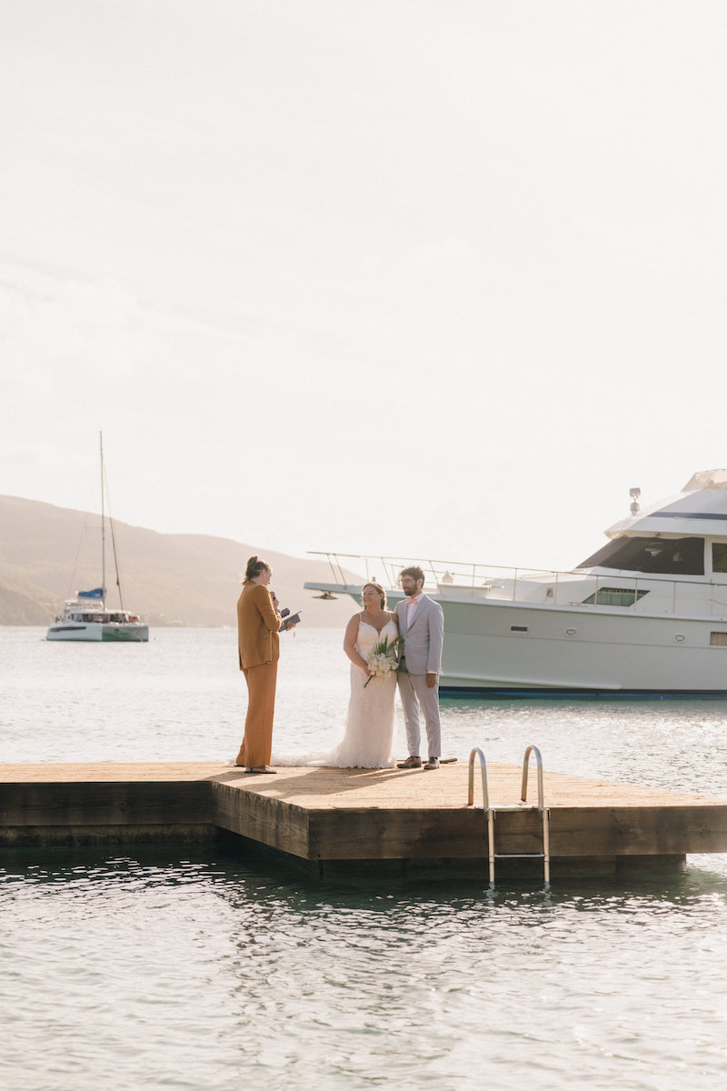 A couple stands with an officiant on a dock by the water, with yachts and sailboats in the background.