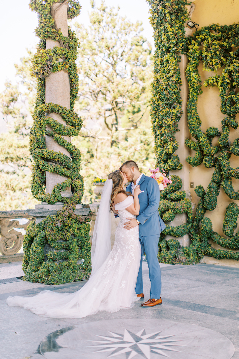 Bride and groom embracing and kissing outdoors in front of ivy-covered columns, with sunlight and greenery in the background.