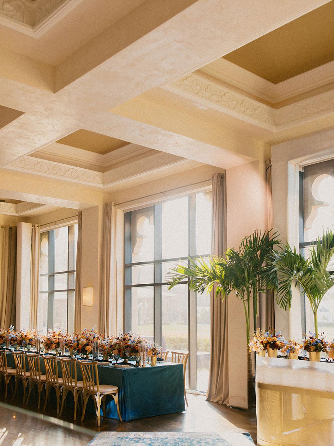 Elegant banquet hall with large windows, long dining table set with florals, gold chairs, and potted palm plants; natural light fills the room.