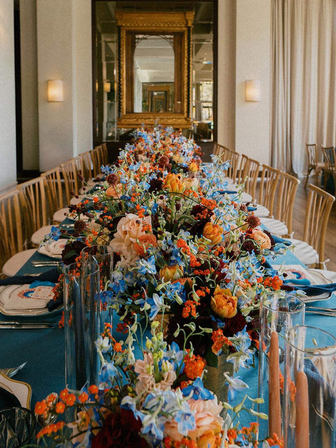 A long dining table set with blue tablecloth, floral centerpieces featuring blue and orange flowers, plates, glassware, and wooden chairs in a modern, elegant room.