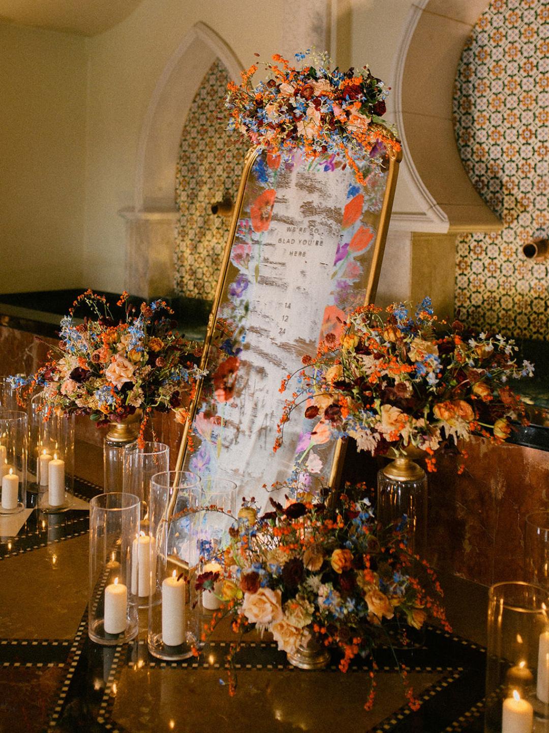 A large mirror decorated with colorful flowers and surrounded by lit candles, with text written on the mirror, displayed in an ornate room with patterned tiles.