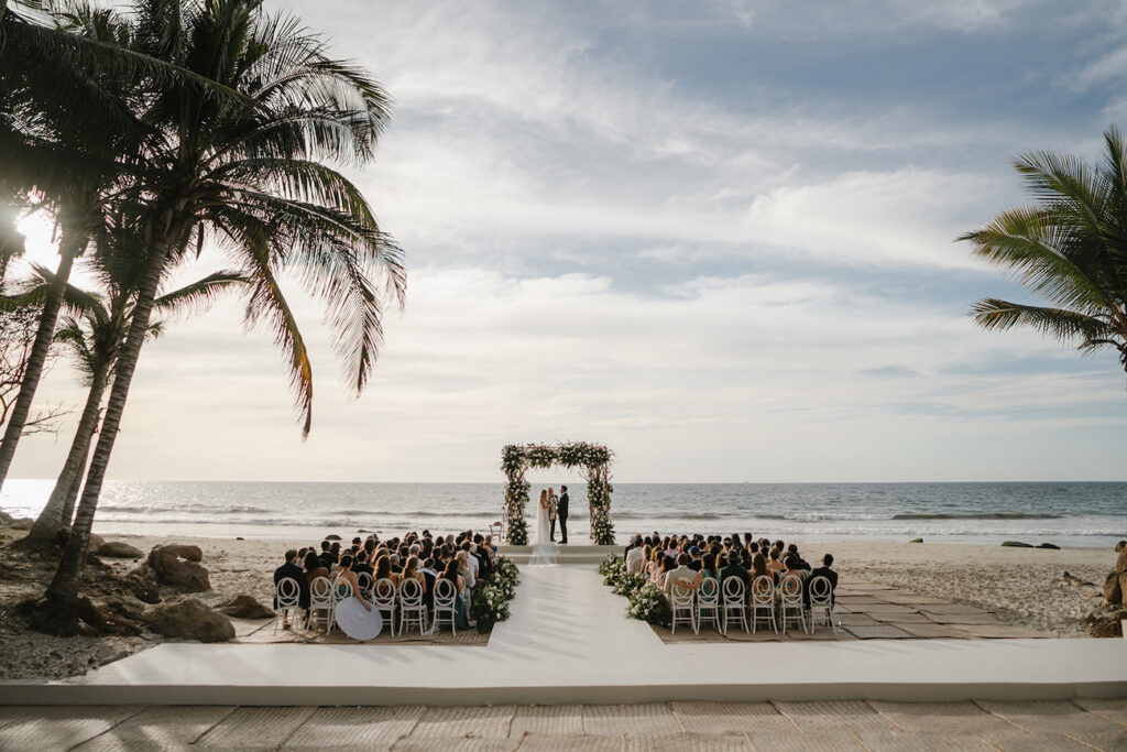 A wedding ceremony takes place on a beach, with guests seated facing an altar decorated with flowers, under palm trees by the ocean.