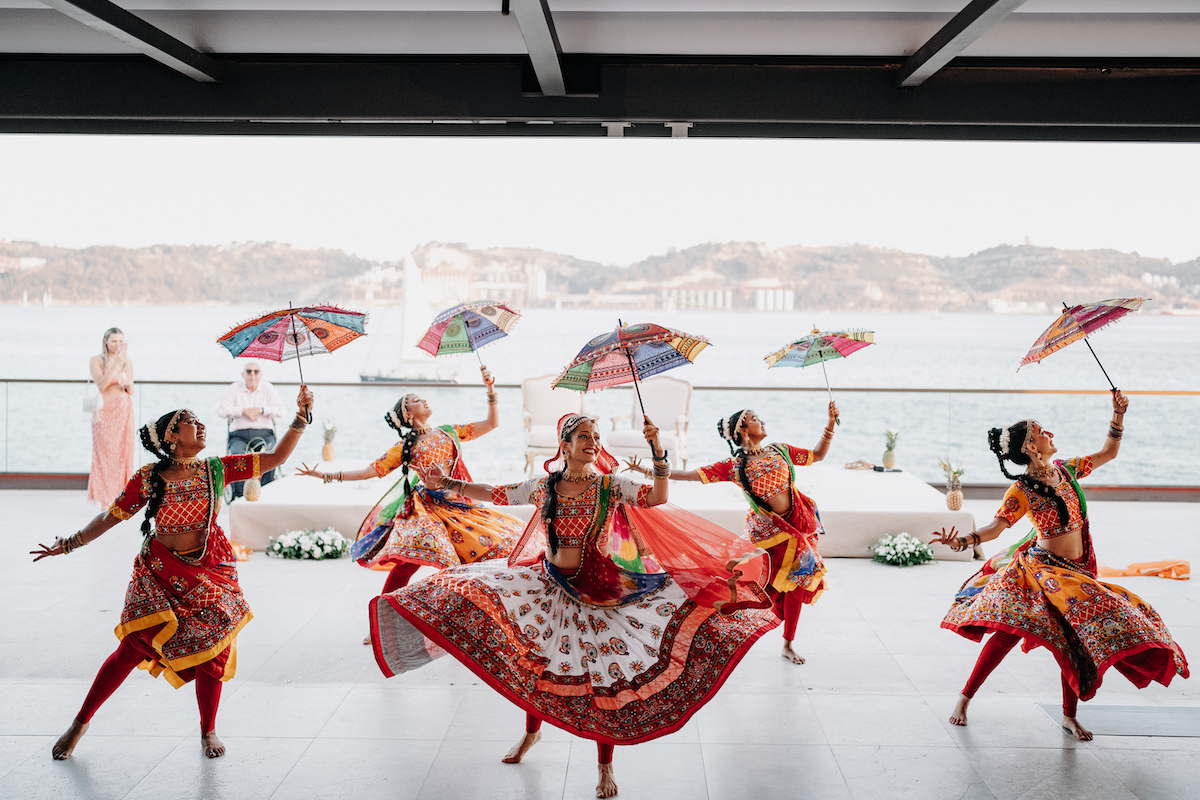 Five dancers in traditional Indian attire perform with colorful umbrellas on a waterfront terrace, with hills and water visible in the background.