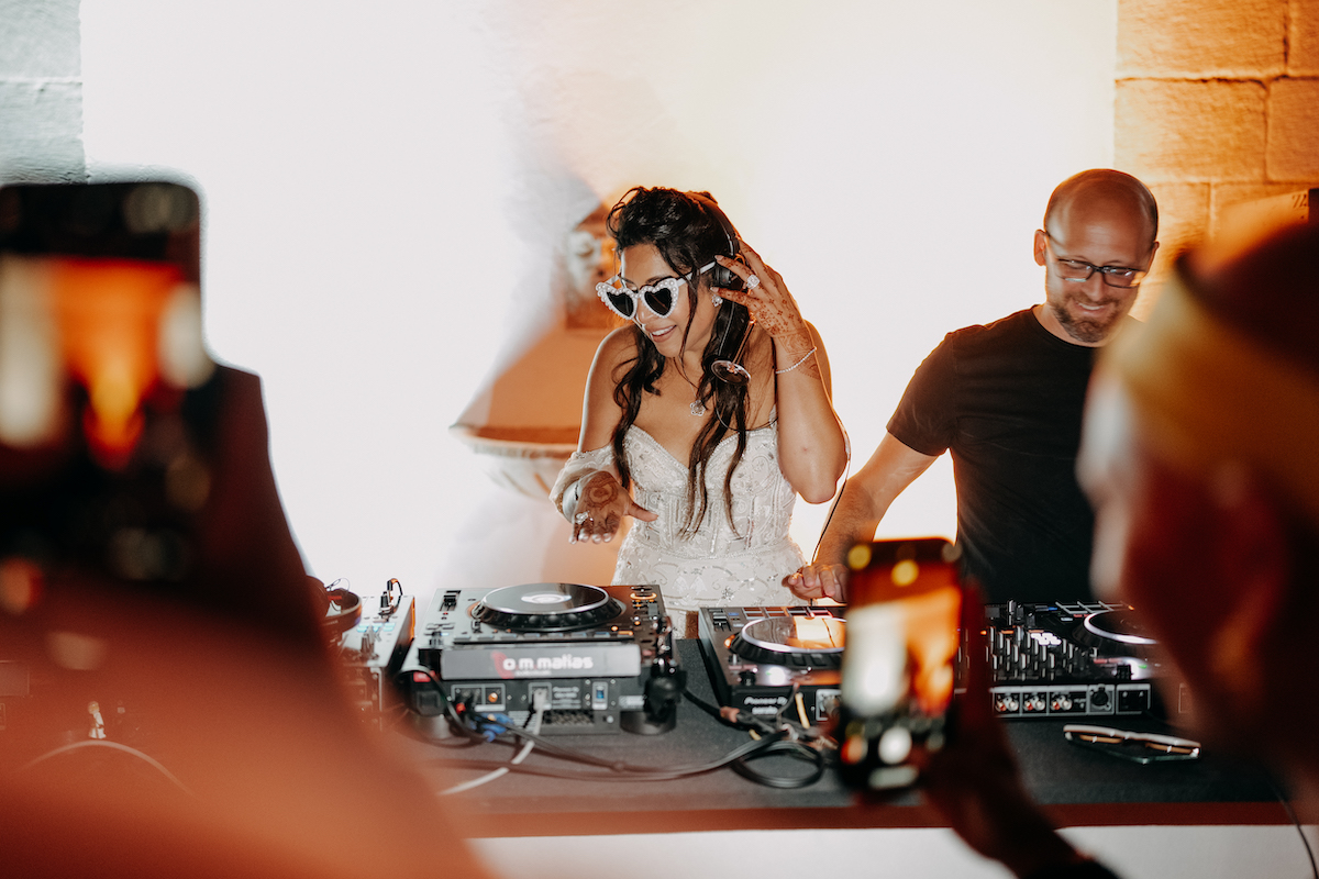 A woman in a white dress and sunglasses DJs next to a man at a mixing console, with people in the foreground taking photos.