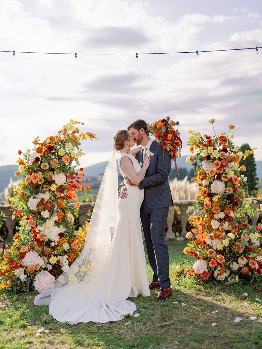 A bride and groom embrace in front of colorful floral arrangements outdoors, with mountains and a cloudy sky in the background.
