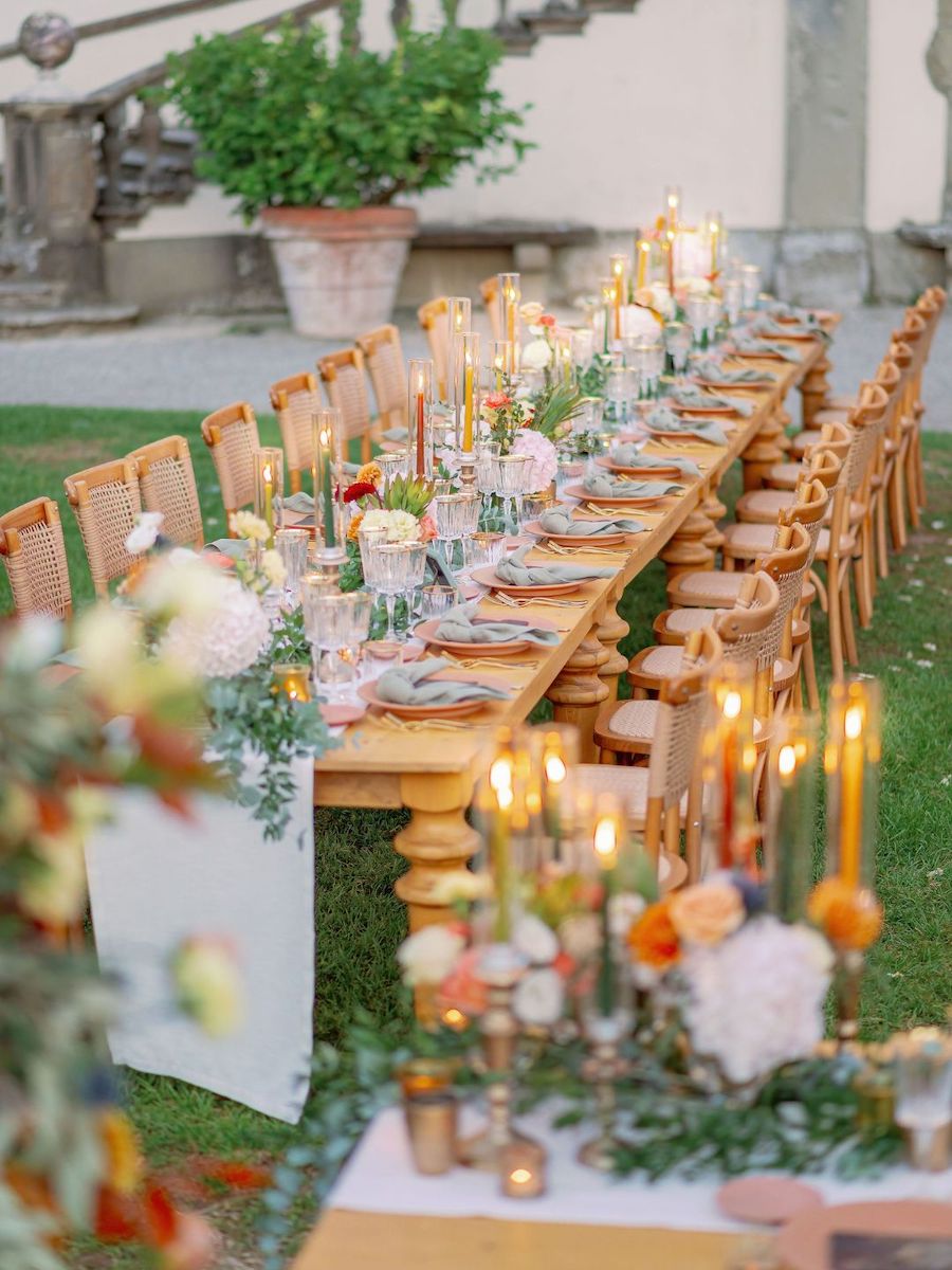 A long outdoor dining table is set with candles, flowers, plates, and glasses, surrounded by wooden chairs on a grassy lawn.