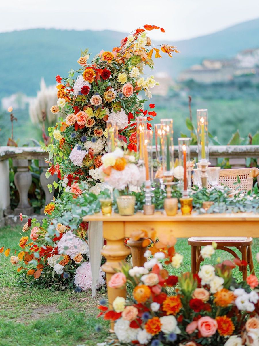 Outdoor table decorated with colorful flowers, candles, and vases, surrounded by floral arrangements, with green hills and stone railing in the background.