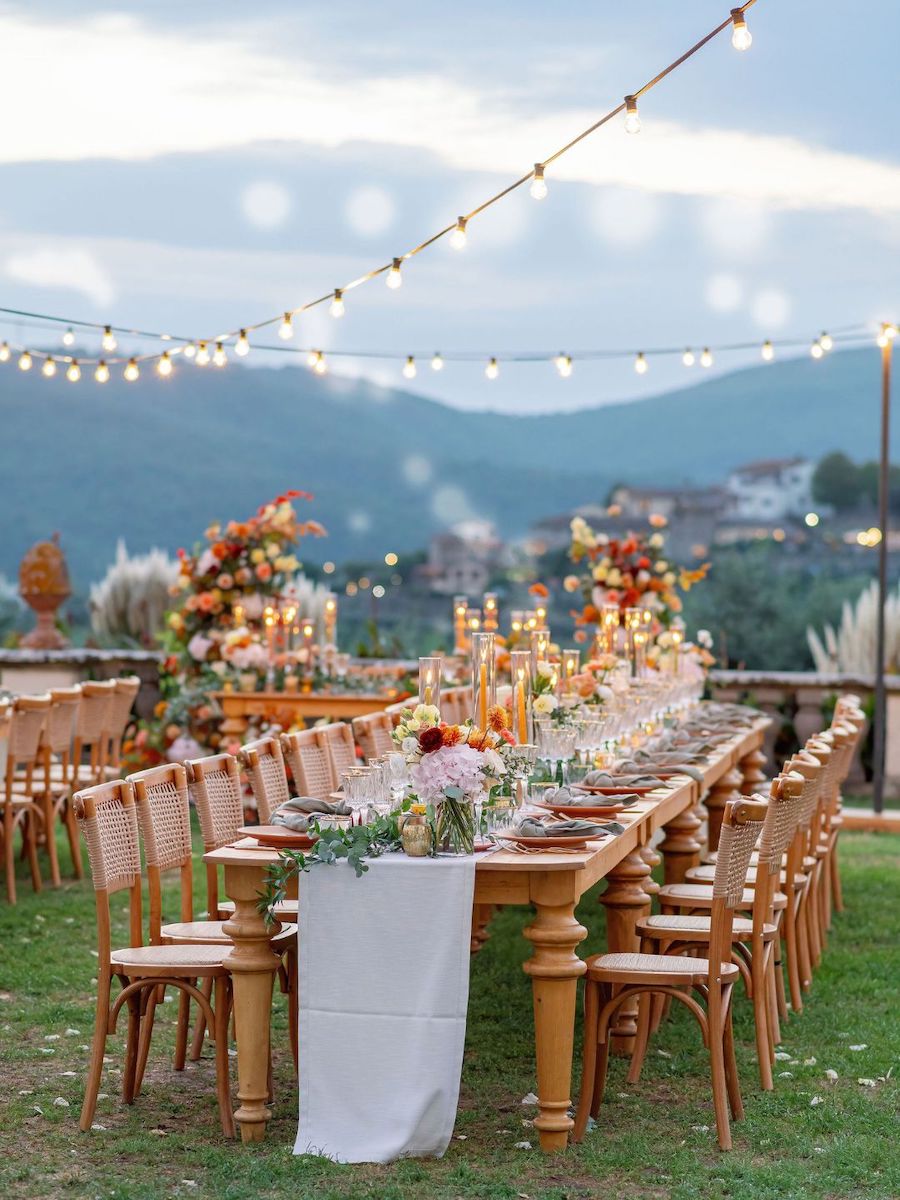 A long outdoor dining table is set with flowers, plates, and candles, surrounded by wooden chairs, under string lights with mountains and buildings in the background.