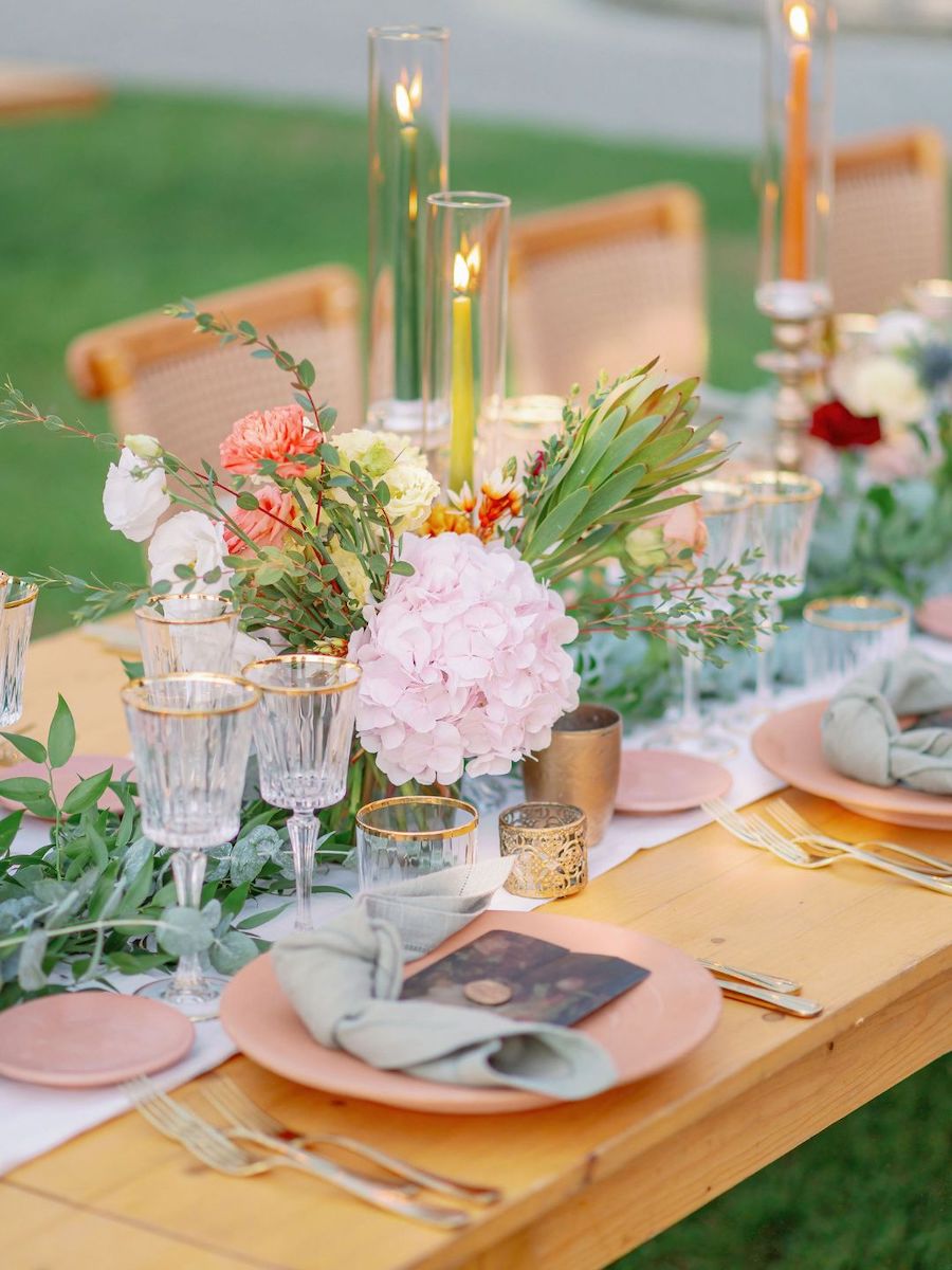 A wooden table set for an outdoor event with glassware, plates, cloth napkins, candles, and a floral centerpiece featuring pink and white flowers and greenery.