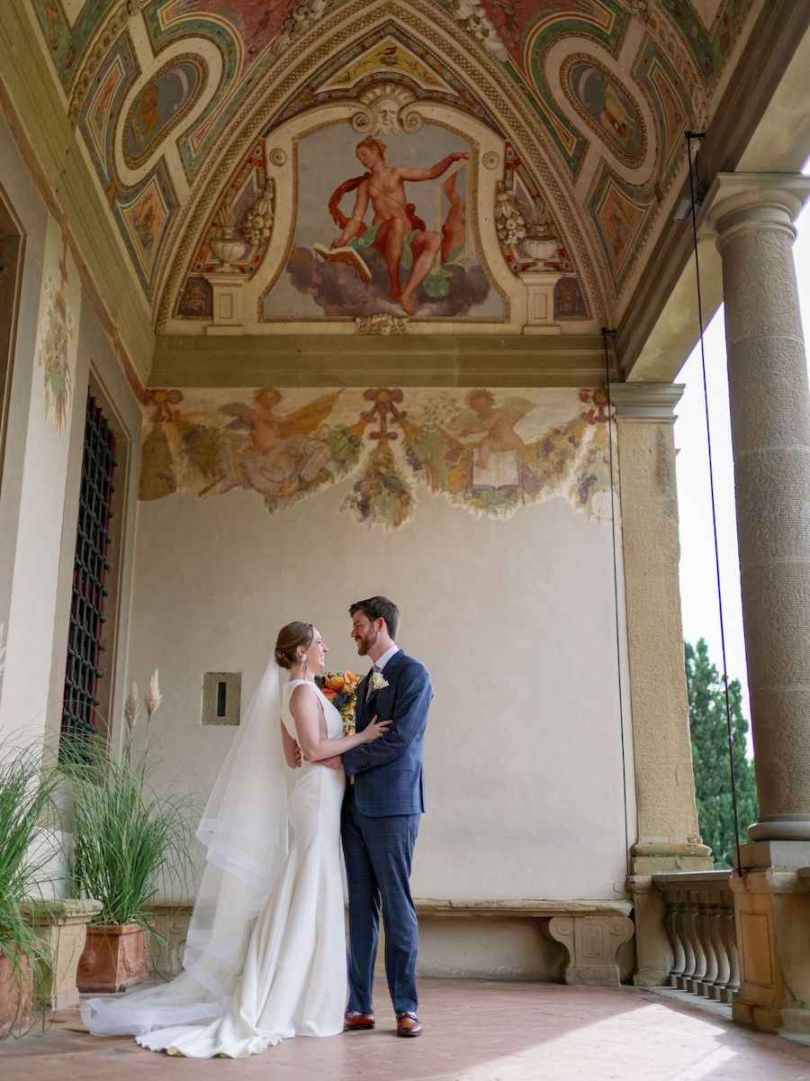 A bride and groom stand facing each other in an ornate, arched hallway with painted ceilings and classical architectural details.