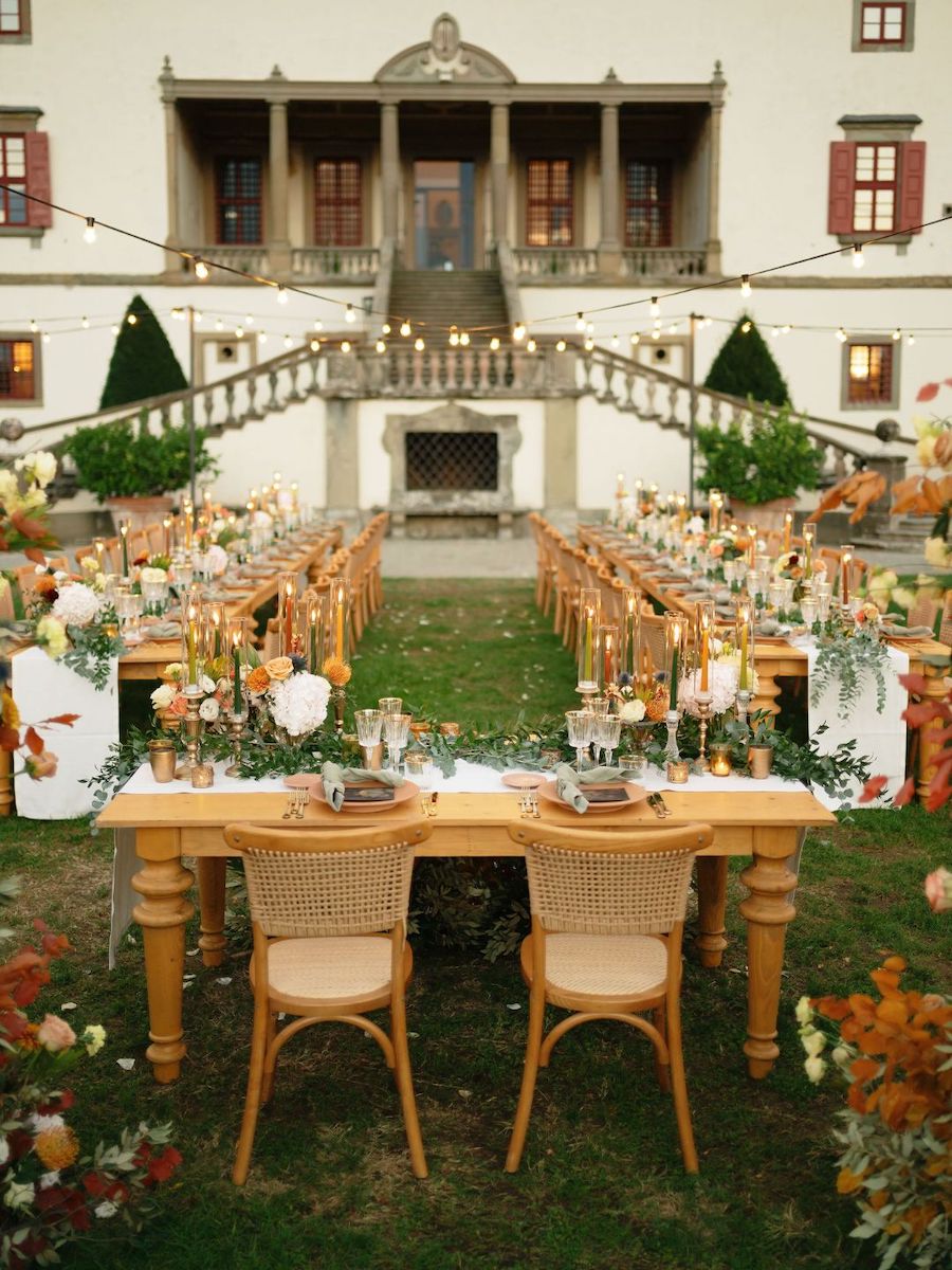 Long banquet tables set for an outdoor event with floral arrangements and string lights, positioned in front of a grand building with stairs and columns.
