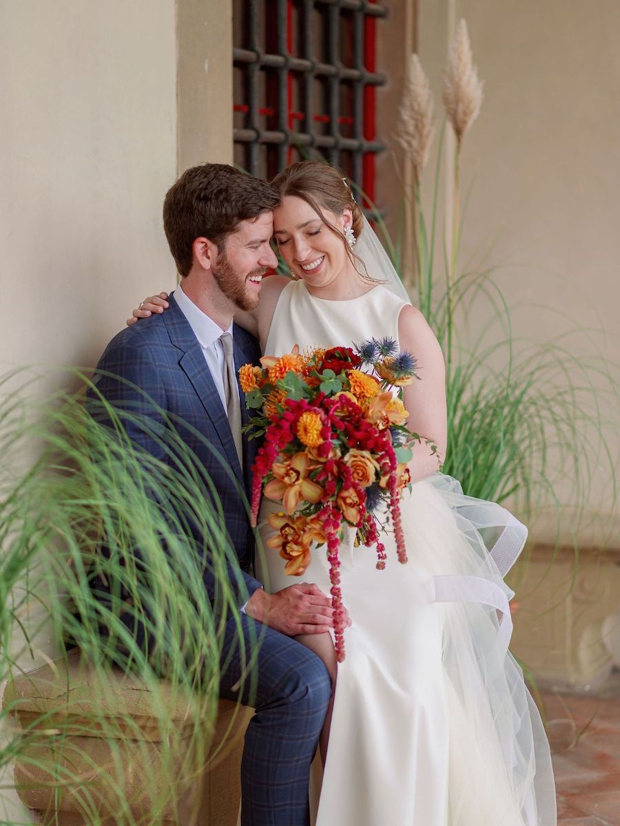 A bride and groom sit closely together, smiling, with the bride holding a colorful bouquet of flowers in an outdoor setting.