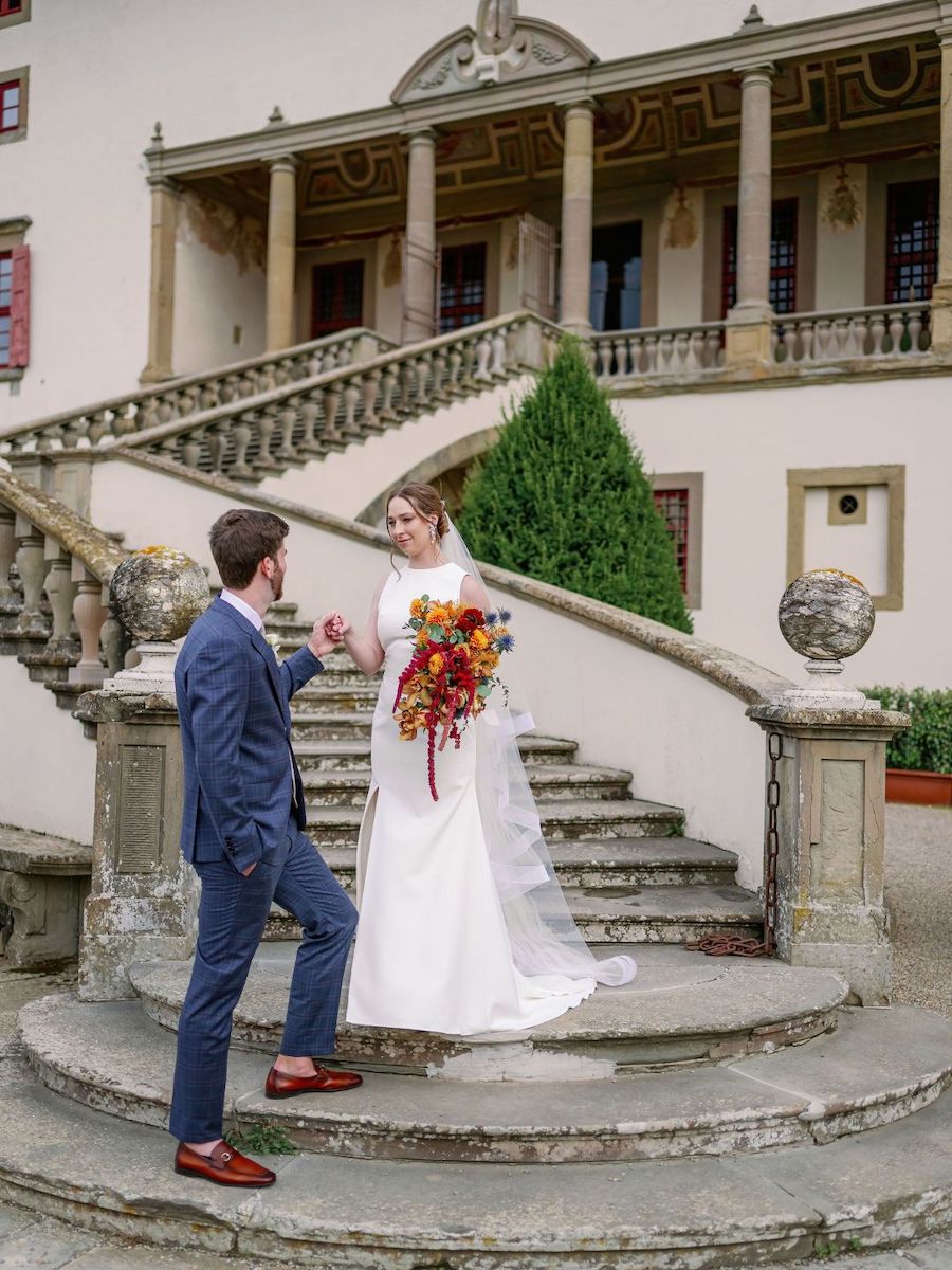 A bride and groom stand on stone steps outside a grand building; the bride holds a bouquet of flowers and they look at each other.