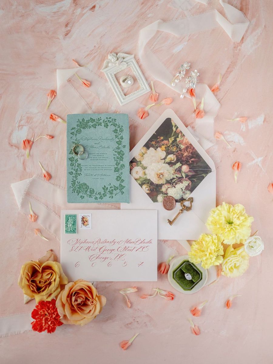 Flat lay of a wedding invitation suite with a teal card, envelope, floral photo, ring in a box, yellow and orange flowers, and scattered flower petals on a pink background.