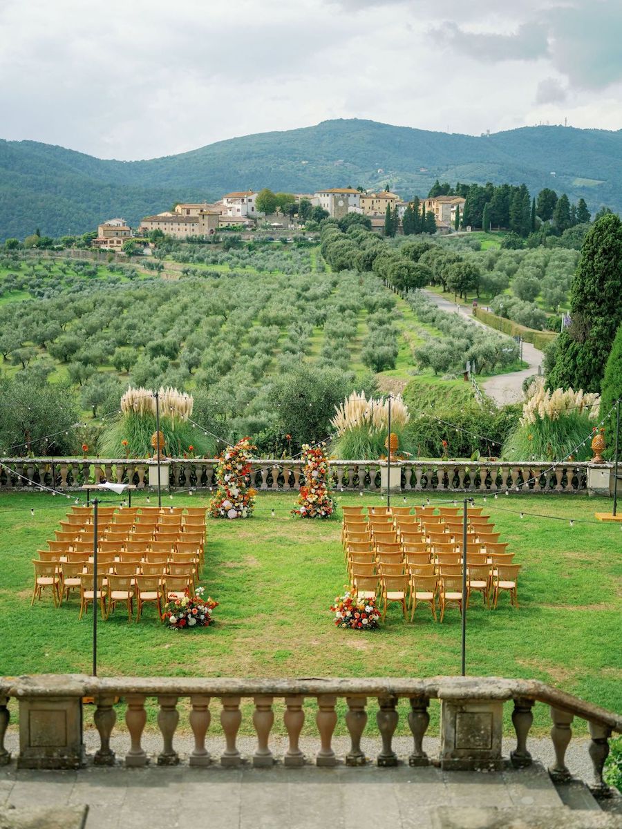 Rows of chairs arranged for an outdoor event on a lawn, with floral decorations, overlooking a scenic hillside village and green landscape.
