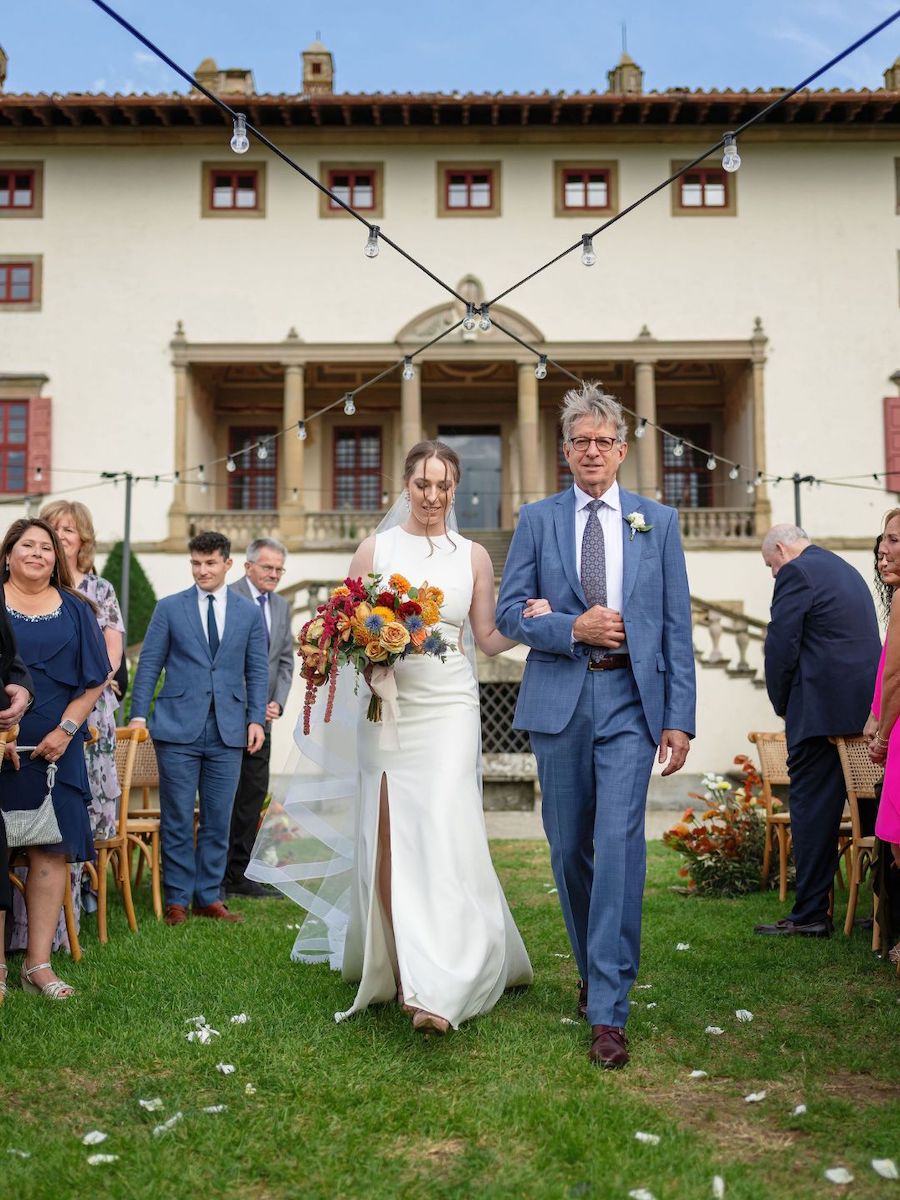 A bride in a white dress and a man in a blue suit walk down an outdoor aisle, surrounded by guests, in front of a large, elegant building.