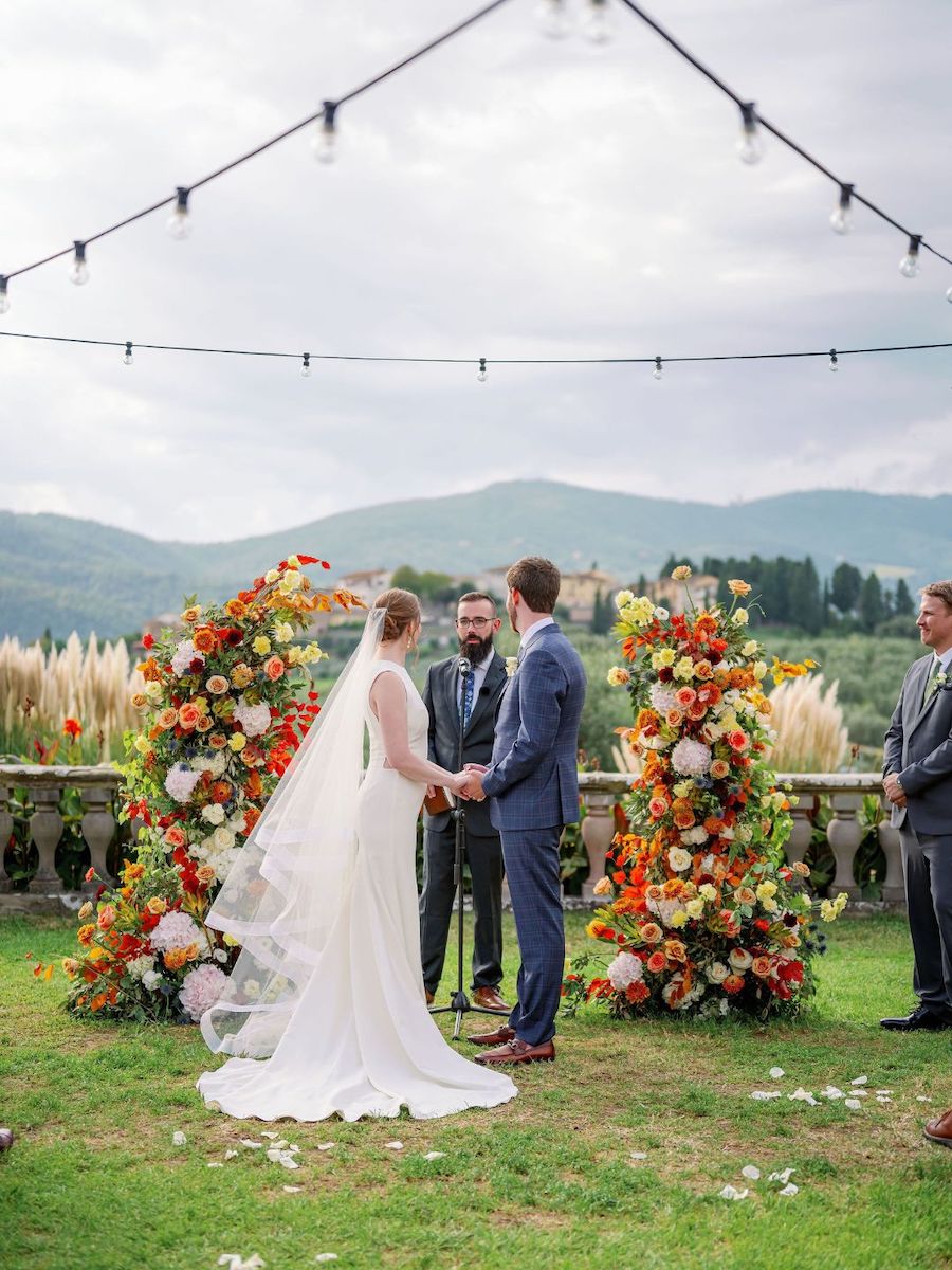 A woman in a white dress with pants underneath sits on a light green dresser next to a vase of colorful flowers and some books.