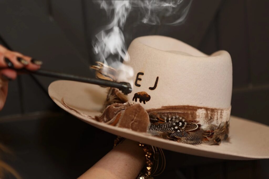 A hand burns initials into a white cowboy hat with feathers and a decorative band, using a smoking branding tool.