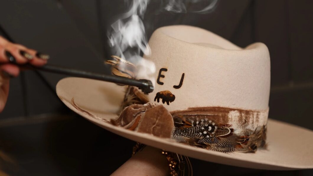 A hand burns initials into a white cowboy hat with feathers and a decorative band, using a smoking branding tool.