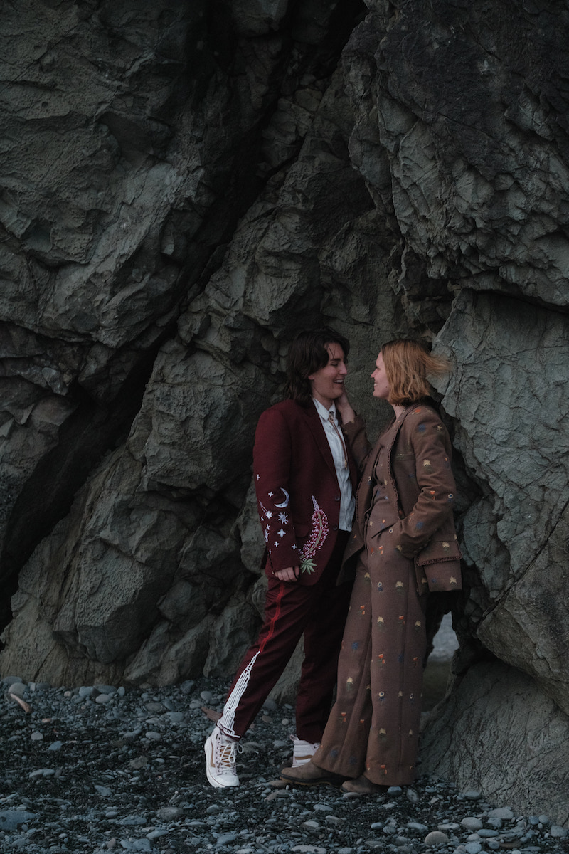 Two people stand facing each other on rocky ground near a large rock formation, wearing patterned suits and coats.