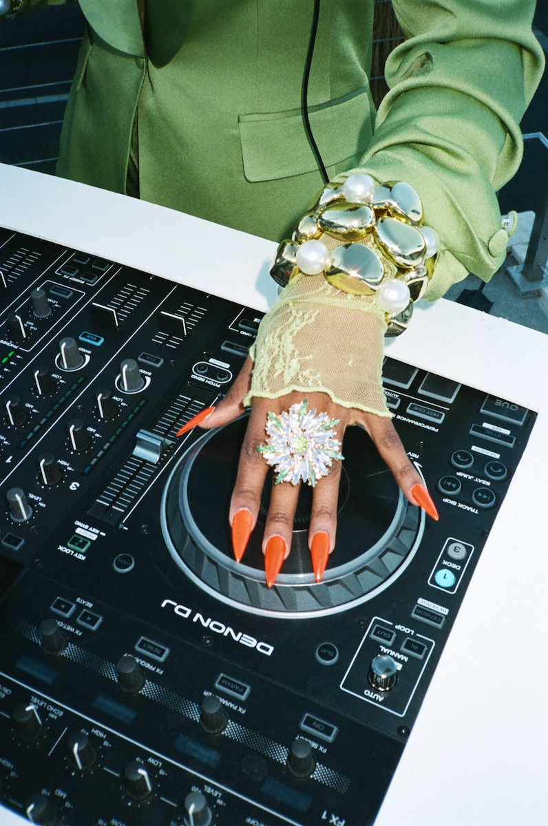 A person wearing a green sleeve, large jewelry, and long orange nails adjusts controls on a Pioneer DJ controller.