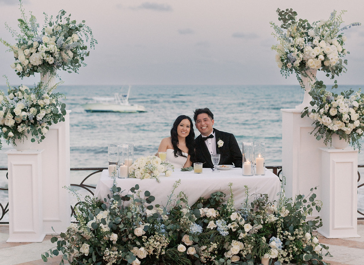 A bride and groom sit together at a flower-adorned table on a seaside terrace, smiling at the camera with the ocean and a boat in the background.