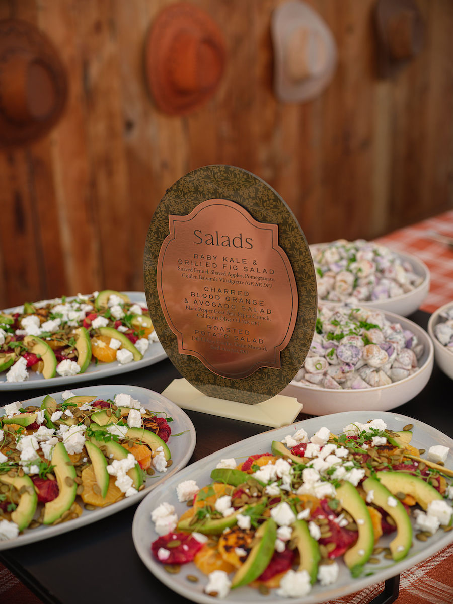 Plates of salad with avocado, feta, and tomatoes are displayed on a table next to a copper-colored sign labeled "Salads" with additional small bowls of salad in the background.