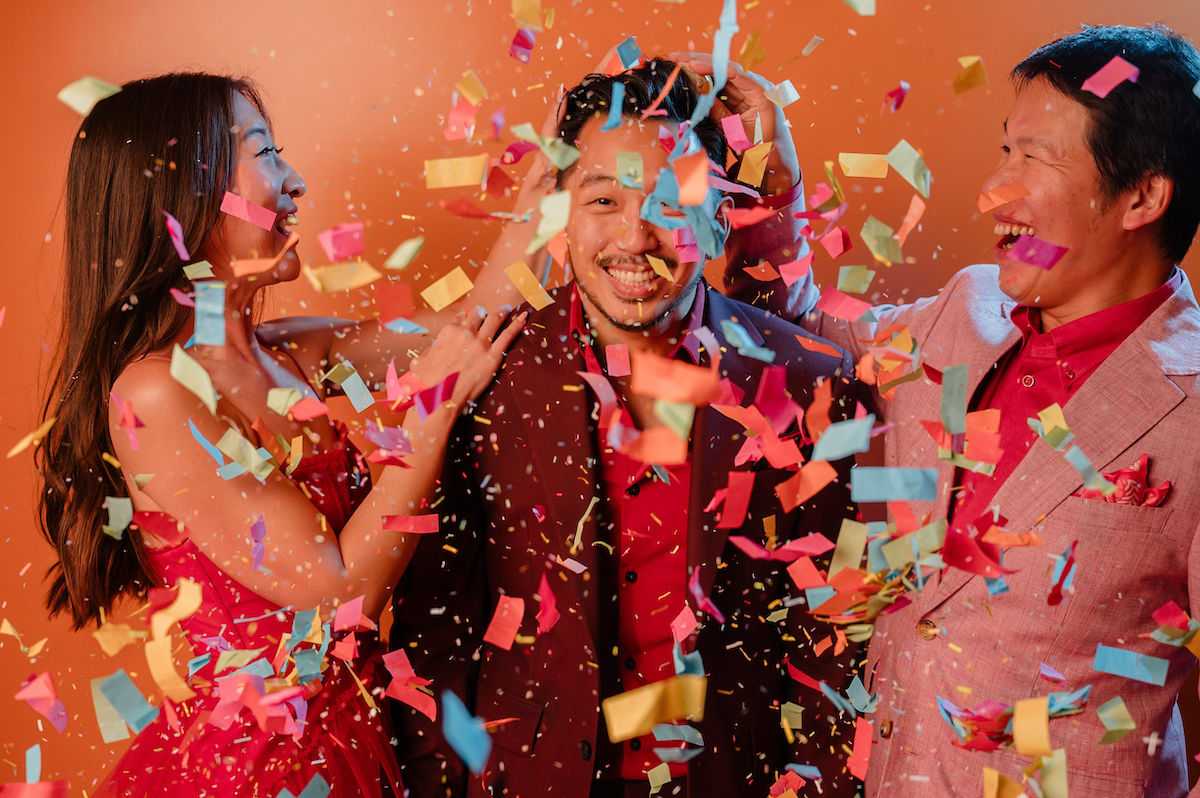 Three people in festive clothing smile and celebrate as colorful confetti falls around them against an orange background.