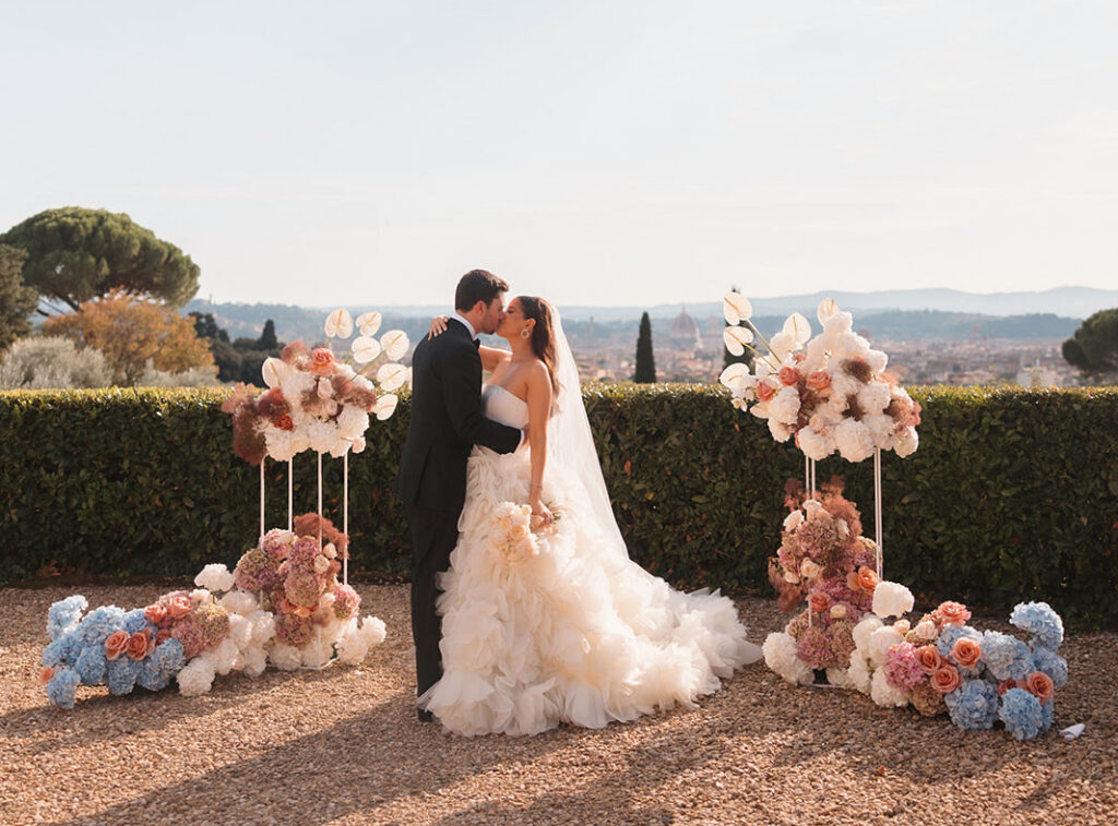 A bride and groom kiss outdoors between two floral arrangements, with a scenic landscape and city in the background.