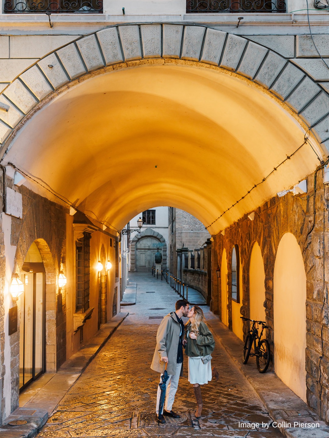A couple stands under a lit stone archway in a narrow, cobblestone alley at dusk, sharing a kiss.