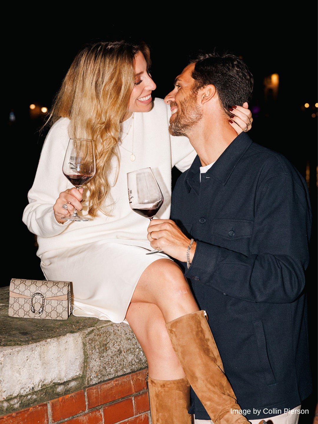 A woman and a man sit together outdoors at night, each holding a glass of red wine. The woman is sitting on a ledge, smiling at the man.