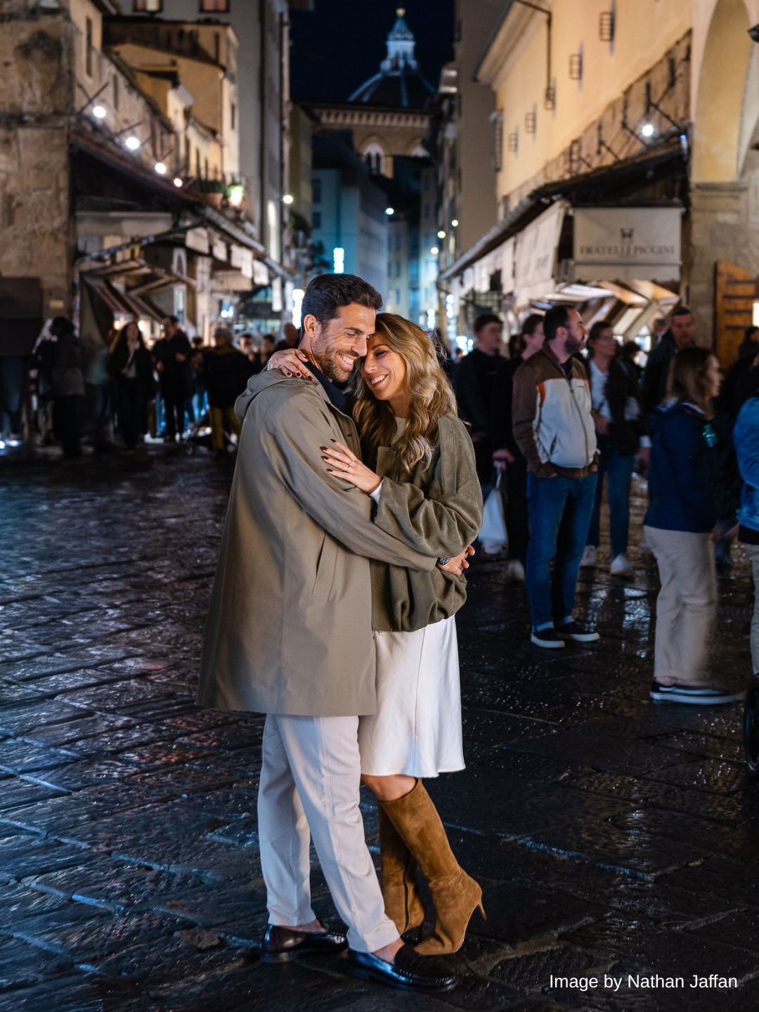 A couple embraces and smiles on a busy, cobblestone street at night, surrounded by people and illuminated shopfronts.