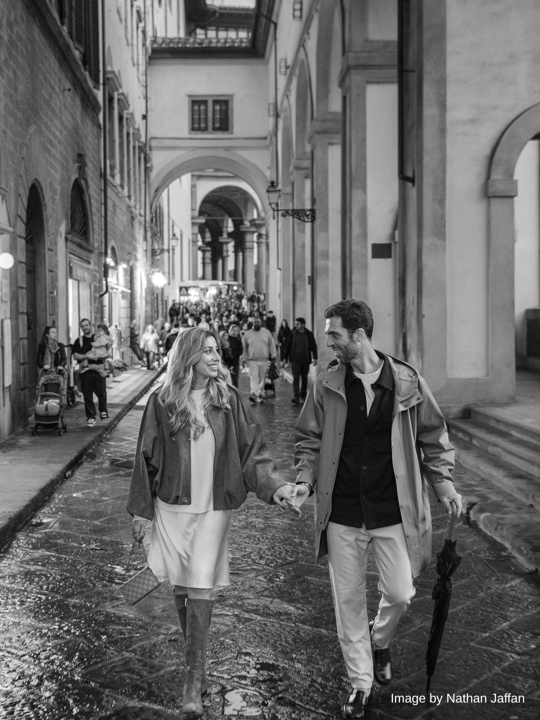 A couple walks hand in hand down a cobblestone street lined with historic buildings, as other people stroll in the background.