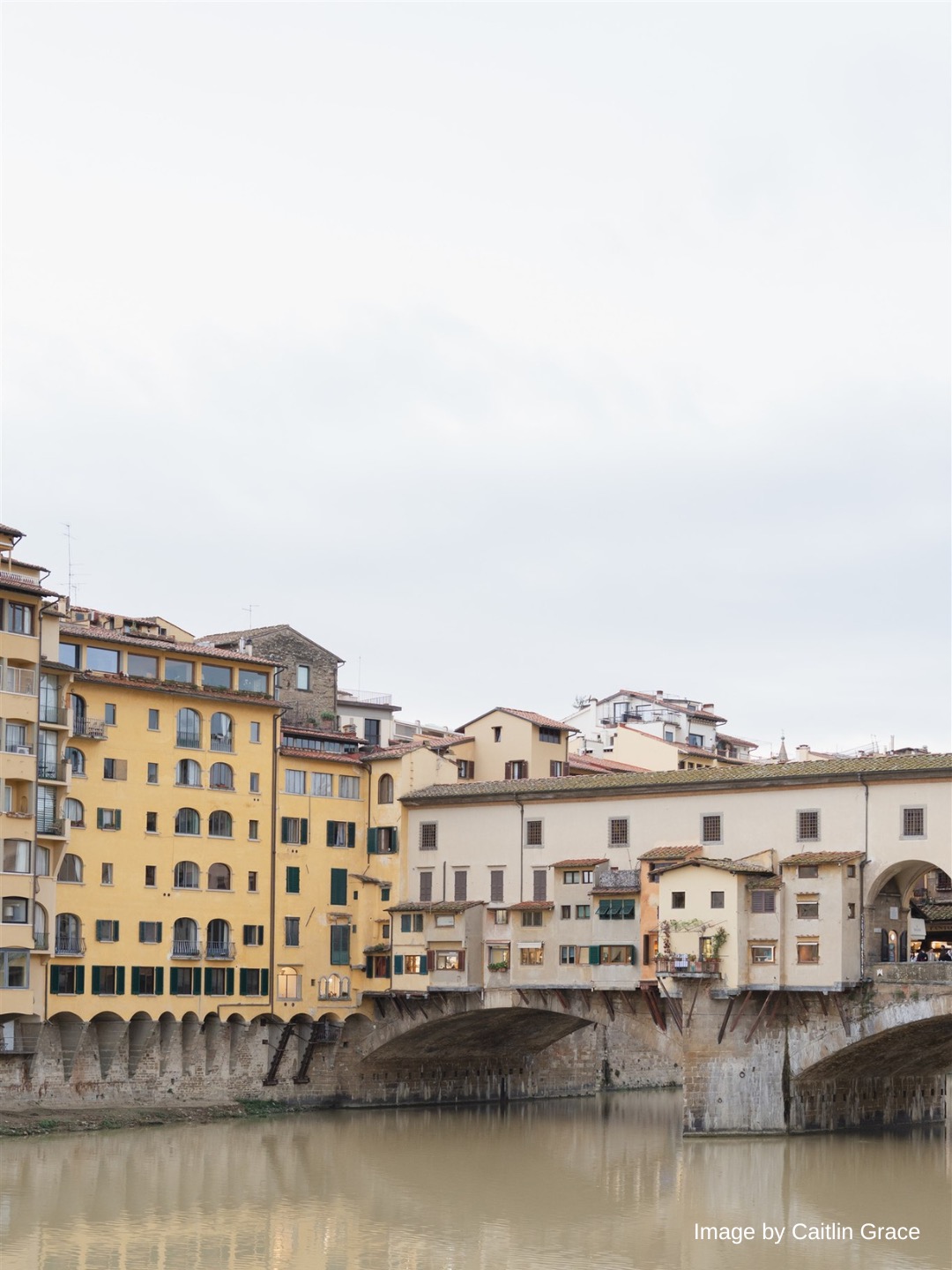 View of the Ponte Vecchio bridge over the Arno River in Florence, with historic yellow and white buildings lining the waterfront under a cloudy sky.