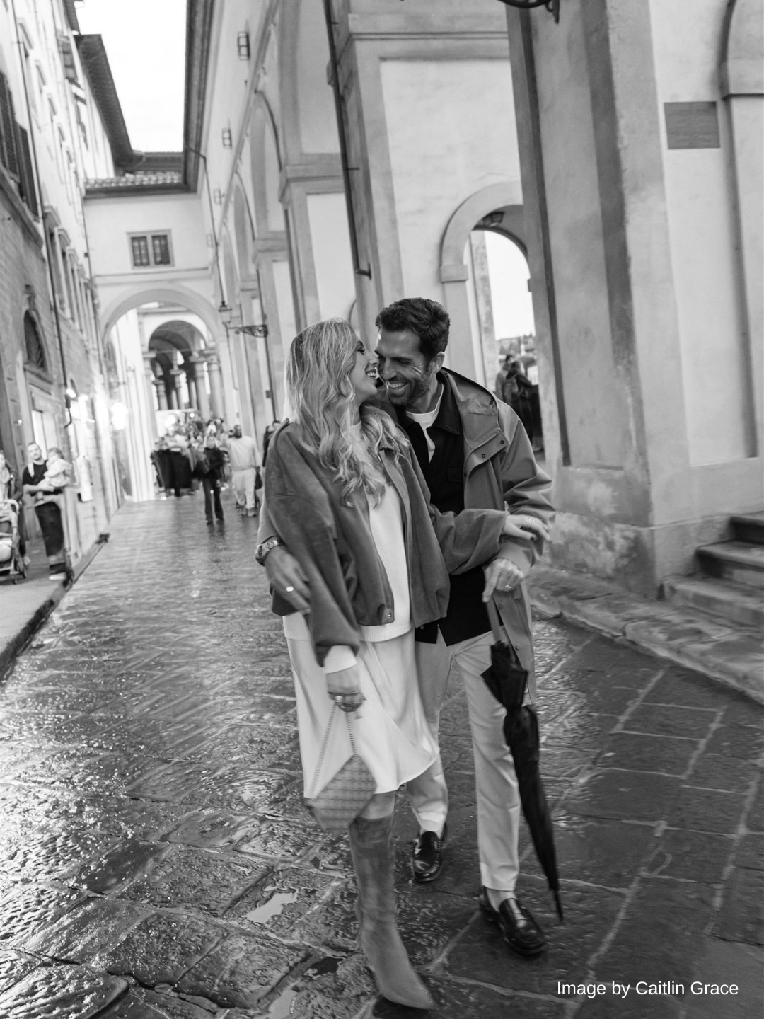 A couple walks closely together on a wet cobblestone street lined with arches and buildings. The man holds an umbrella and both appear to be smiling. The scene is in black and white.