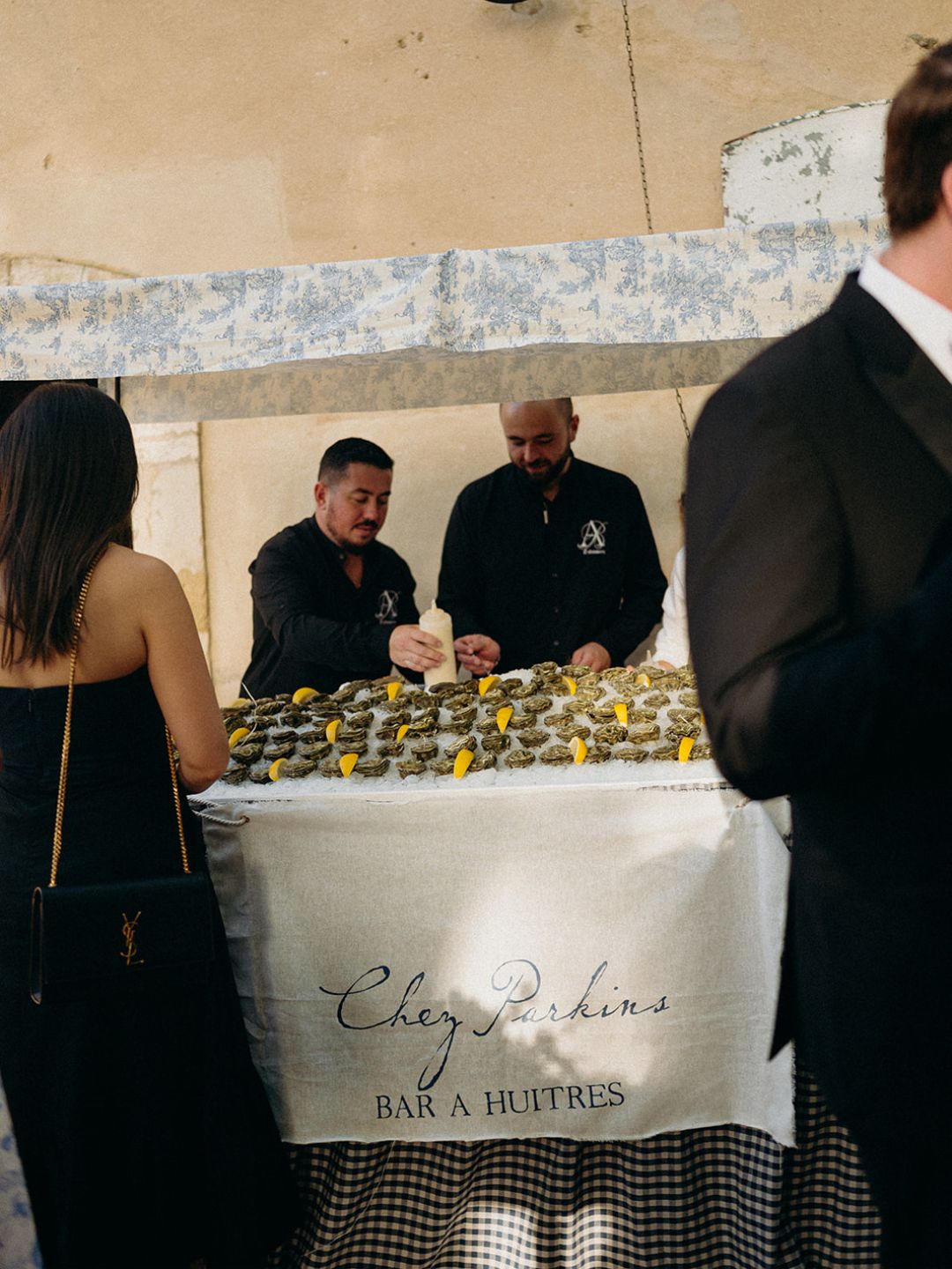 Two men shuck oysters behind a table covered with oysters and lemon wedges at a "Chez Fardins Bar à Huîtres" stand during an event.