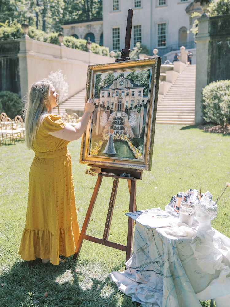 A woman in a yellow dress paints a large outdoor scene on an easel in a garden, with art supplies on a table nearby.