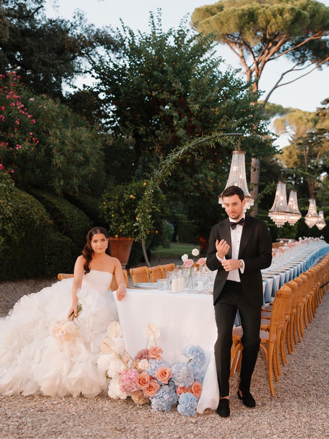A bride and groom pose by a long, elegantly decorated outdoor table with floral arrangements and hanging chandeliers in a garden setting.